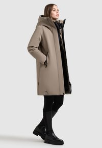 Beige parka střední délky s kapucí, s černými detaily a velkými kapsami. Tkanina vypadá hladce, s měkkou podšívkou.