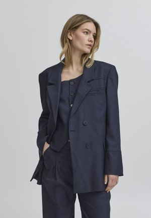 Námorně modrý přizpůsobený blazer s volným střihem, dvojitými knoflíky a strukturovaným designem ramen, spárovaný s odpovídajícím topem a kalhotami.