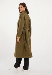 Long manteau vert olive en tissu texturé, doté d'une taille ceinturée, d'un grand col et de poignets à boutons, associé à un pantalon noir et à des baskets blanches.