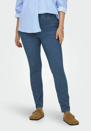 Skinny jeans in dark blue denim with a smooth texture. Feature a mid-rise waist and subtle stitching. Paired with tan clogs and a blue striped shirt.