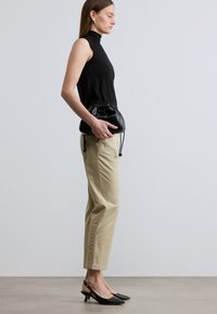 Femme en pantalon beige, col roulé sans manches noir et talons noirs tenant un petit sac à cordon en cuir noir sur un fond uni.