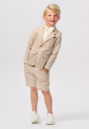 Blazer beige avec deux poches, associé à un short assorti. Chemise blanche en dessous et un nœud papillon. Des baskets blanches complètent l'ensemble.