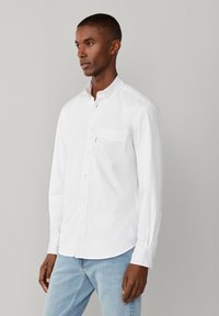 Homme portant une chemise blanche à boutons avec une poche poitrine et un jean bleu clair, debout devant un fond gris clair uni.