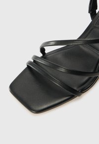 Sandal cu toc pătrat, din piele neagră, cu mai multe curele subțiri ce traversează partea de sus a piciorului și o suprafață texturată netedă.