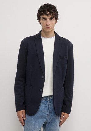 Giovane uomo con capelli corti e ricci, che indossa un blazer blu navy sopra una camicia bianca strutturata e jeans azzurri chiari, su uno sfondo neutro.