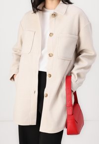Beige wool-blend shirt jacket with two chest pockets and large brown buttons, worn over a white shirt and paired with a red handbag.