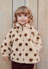 Veste en fausse fourrure beige à pois marron, fermeture éclair sur le devant et col montant. Texture douce, avec manches longues et coupe décontractée.