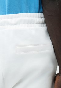 Κλείσιμο σε σκούρο δέρμα ενός μπράτσου δίπλα σε λευκό παντελόνι με ελαστική μέση και οριζόντια πίσω τσέπη, και μπλε πουκάμισο από δίχτυ περασμένο μέσα.