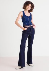 Top corto in denim con chiusura a bottoni, abbinato a jeans svasati scuri. La modella indossa stivaletti alla caviglia chiari. Design semplice e minimalista.
