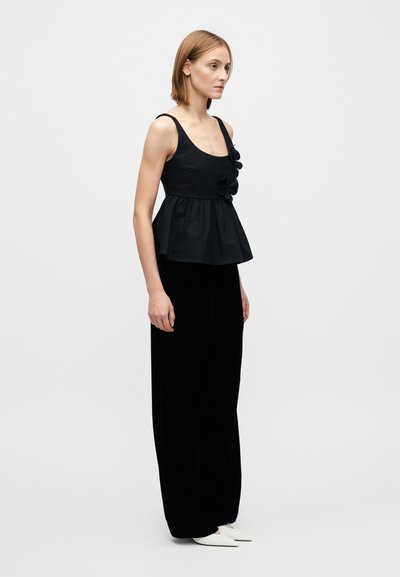 Svart peplum-topp med volangdetaljer, kombinert med svarte vide bukseben og hvite spisse flate sko. Glatt stofftekstur og tettsittende design.