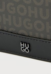 Accessoire en cuir noir présentant un motif « HUGO » embossé, une texture lisse et un logo métallique argenté, monté sur un fond épuré.