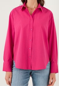 Camicia lunga a maniche con bottoni di colore rosa in un tessuto leggero, dotata di colletto classico e vestibilità rilassata, abbinata a jeans azzurri chiari.