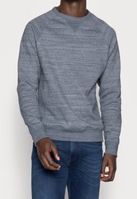 Felpa grigia con collo tondo, tessuto strutturato, maniche raglan e polsini a coste. Abbinata a jeans blu, mostrata frontalmente.