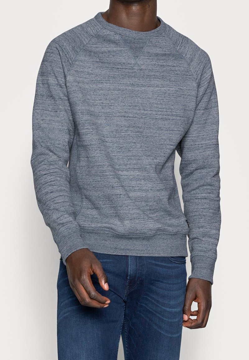 Sudadera gris de cuello redondo con tejido texturizado, mangas raglán y puños acanalados. Combinada con jeans azules, exhibida de frente.