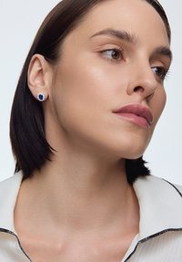 Złote kolczyki w kształcie stóp z centralnym niebieskim kamieniem, otoczonym małymi przezroczystymi kamieniami. Projekt ma okrągły kształt i polerowane wykończenie.