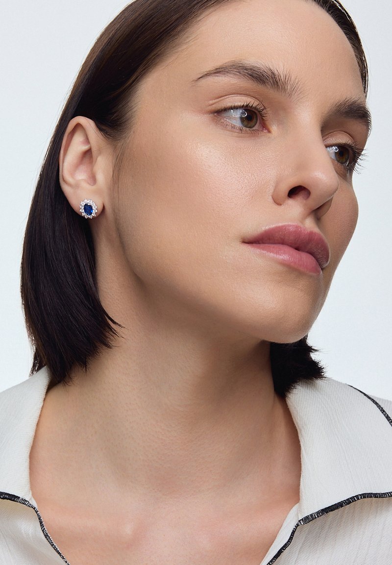 Złote kolczyki w kształcie stóp z centralnym niebieskim kamieniem, otoczonym małymi przezroczystymi kamieniami. Projekt ma okrągły kształt i polerowane wykończenie.