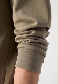 Long-sleeve shirt cuff in beige fabric, featuring a gathered texture with a ribbed hem. Smooth and soft finish.