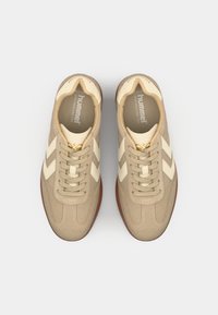 Beige sneakers with a textured suede upper, white leather accents, and a rubber sole. Features a classic lace-up design and logo on the tongue.