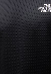 Svart teksturert stoff med små hevede prikker, med en hvit logo av "The North Face" øverst til venstre.
