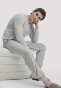 Graues Fleece-Loungewear-Set mit einem Rundhalsausschnitt und eng zulaufenden Hosen. Verfügt über eine glatte Textur und eine entspannte Passform. Modell sitzt auf einer strukturierten weißen Oberfläche.