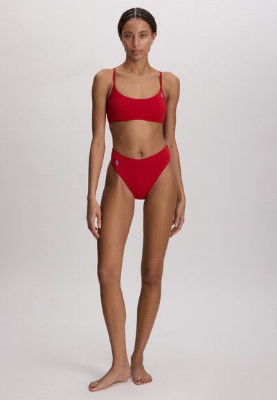 Λεπτή γυναίκα που στέκεται ξυπόλητη φορώντας κόκκινο μαγιό δύο τεμαχίων με λεπτές τιράντες και λογότυπα της μάρκας στο στήθος και το ισχίο, σε απλό φόντο.