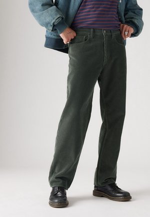 Person trägt dunkelgrüne Cordhose, schwarze Lederschuhe, ein gestreiftes Hemd und eine blaue Jeansjacke mit Händen nahe den Taschen.