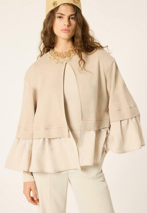 Mujer con chaqueta beige de capas con mangas con volantes, pantalones crema, collar decorativo dorado y sombrero con textura a juego sobre fondo liso.