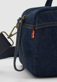 Bolso de mezclilla azul con forma cuadrada, costuras amarillas, un asa superior y una correa desmontable. Presenta una pequeña etiqueta roja de Levi's.