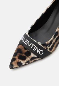 Chaussure plate à bout pointu en tissu imprimé léopard avec une bande élastique noire sur le devant affichant le texte VALENTINO en blanc.