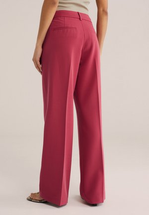 Osoba oblečená v širokých fialových nohaviciach s vysokým pásom a béžovom tielku bez rukávov, pohľad zozadu od pásu po nohy.