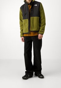 Fleecevest in groen en zwart met ritssluiting, twee borstzakken, hoge kraag en zwarte broek. Model draagt zwarte sneakers.