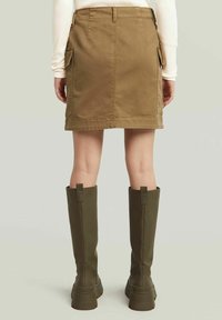 Gonna di denim khaki con tasche laterali, lunghezza media fino alla coscia, abbinata a stivali al ginocchio verde oliva con suola spessa e dettagli con cinturino.