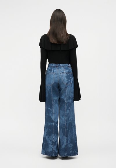 Γυναίκα που φαίνεται από πίσω, φοράει μαύρο μακρυμάνικο τοπ με φαρδιά μανσέτα και μπλε τζιν παντελόνι υψηλής μέσης με φαρδιά πόδια και μπογιατιστό μοτίβο.