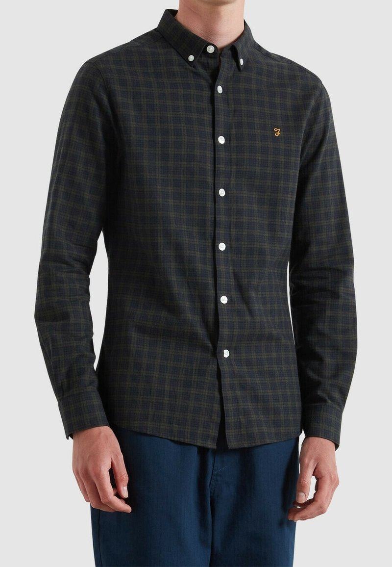 Homme portant une chemise à carreaux vert foncé et noir avec des boutons blancs et un logo brodé sur la poitrine, associée à un pantalon bleu.