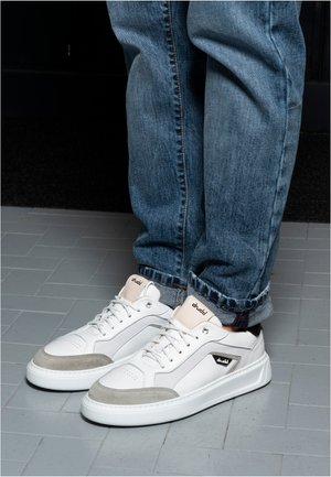 Persoon die een blauwe spijkerbroek en witte sneakers met het label "drudd" draagt, staande op een grijs betegelde vloer.