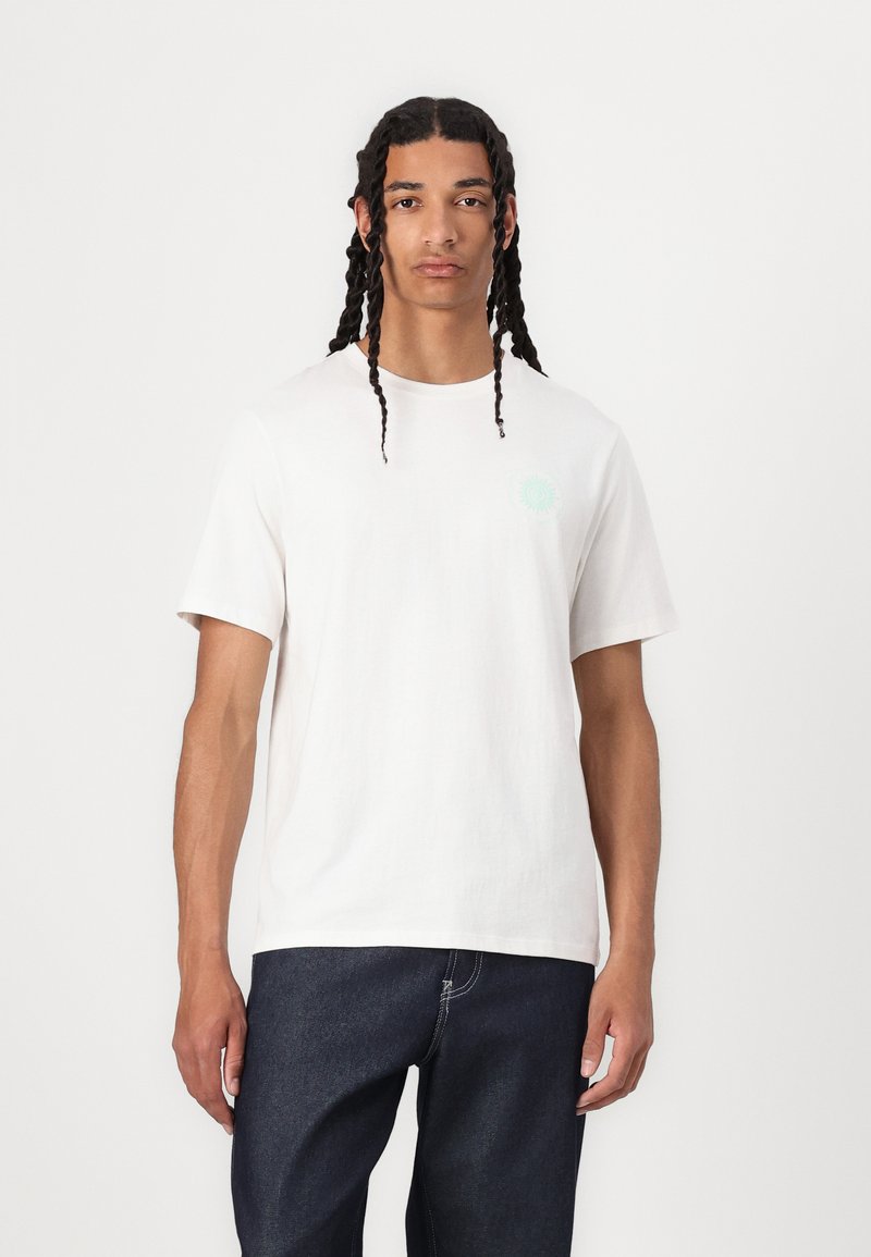 Hvid bomulds T-shirt med korte ærmer, rund hals, der har et lille mintgrønt solmotiv på venstre bryst. Sammen med mørke jeans.
