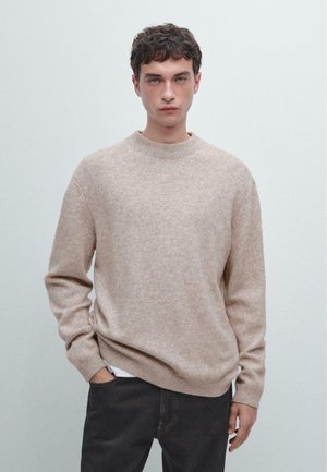 Maglione beige lavorato a maglia con scollatura rotonda, maniche lunghe e orlo a coste. Indossato sopra jeans scuri, con una trama liscia e variazioni sottili.