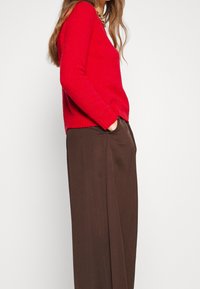 Punainen neulepusero V-pääntiellä ja resorikahdilla, yhdistetty löysään ruskeisiin leveälahkeisiin housuihin, joissa on sivutaskut ja sileä pinta.