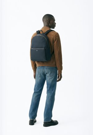 Muž v hnedej bunde, modrých džínsoch, čiernych topánkach a s čiernym ruksakom, stojaci bokom na bielom pozadí.