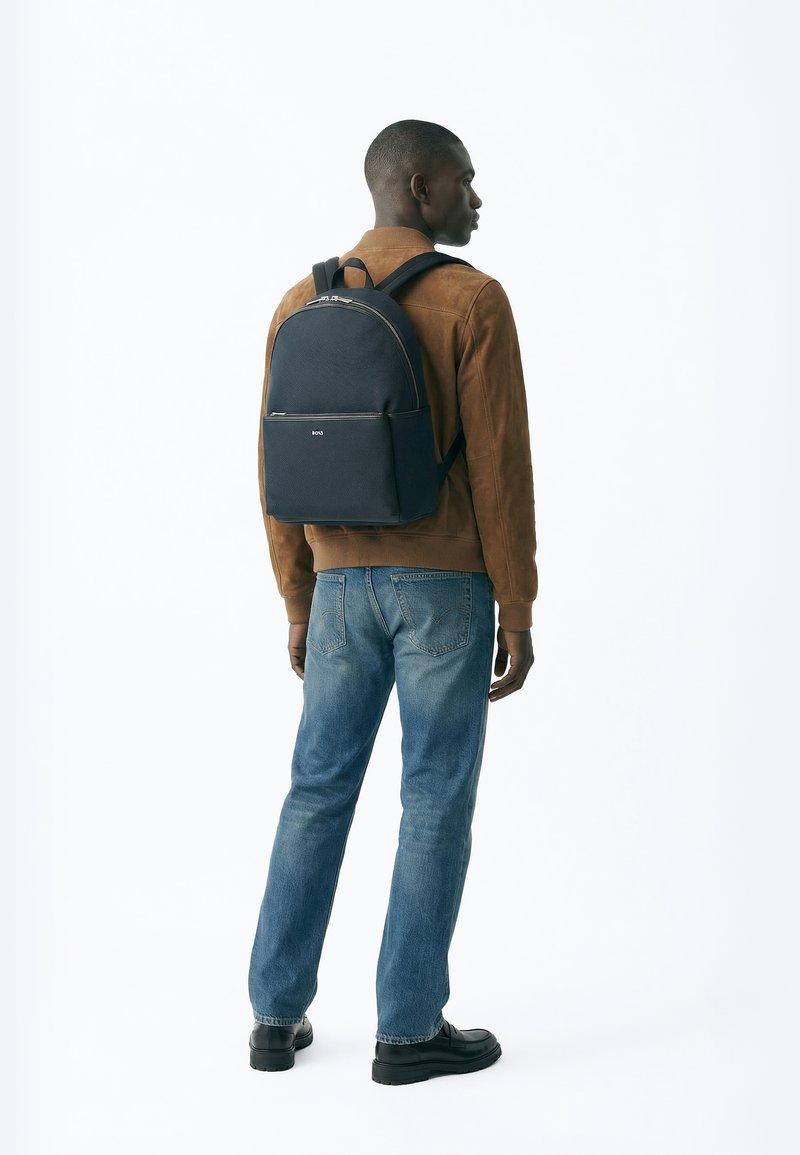 Mann trägt eine braune Jacke, blaue Jeans, schwarze Schuhe und einen schwarzen Rucksack, steht seitlich gewandt vor einem schlichten weißen Hintergrund.