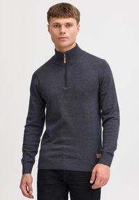 Pull en tricot gris foncé avec un col montant, fermeture éclair quart de zip, poignets et ceinture côtelés, avec un petit patch logo à l'ourlet.