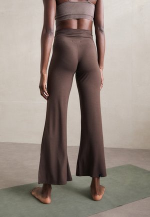 Persoană stând desculță pe un covor de yoga verde, purtând pantaloni largi maro și un top scurt asortat, văzută din spate.
