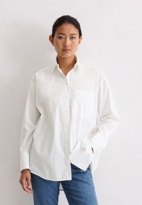 Frau mit dunklem Haar in einem weißen Hemd mit Knöpfen und einer Brusttasche, die vor einer schlichten hellen Wand steht und blaue Jeans trägt.