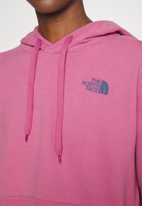 Sudadera con capucha rosa que presenta un bolsillo tipo canguro en la parte frontal, capucha con cordón ajustable y el logo de The North Face en azul marino en el pecho izquierdo.