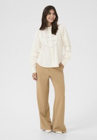 Camicia bianca con dettagli in pizzo e maniche a sbuffo, abbinata a pantaloni beige a gamba larga e décolleté bianche appuntite.