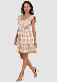 Vestido a cuadros en amarillo pastel, rosa y lavanda. Presenta mangas con volantes, un escote cuadrado y un dobladillo en capas. Llevado con sandalias de tacón transparente.