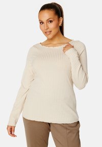 Roomy crème ribbed top met lange mouwen, een golvende halslijn en subtiele textuur. Gecombineerd met lichtbruine broeken, die een ontspannen pasvorm accentueren.