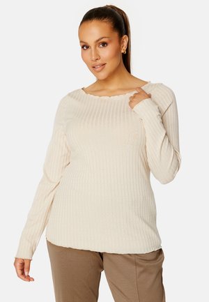 Roomy crème ribbed top met lange mouwen, een golvende halslijn en subtiele textuur. Gecombineerd met lichtbruine broeken, die een ontspannen pasvorm accentueren.