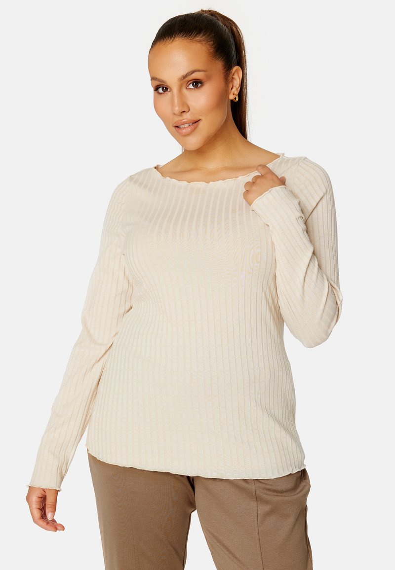 Roomy crème ribbed top met lange mouwen, een golvende halslijn en subtiele textuur. Gecombineerd met lichtbruine broeken, die een ontspannen pasvorm accentueren.
