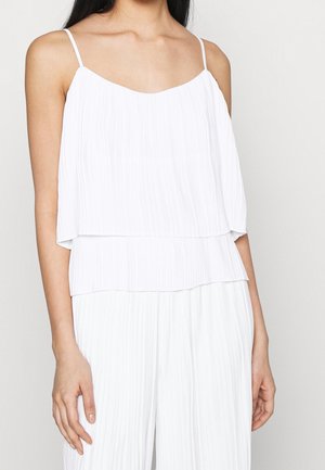 Frau trägt ein ärmelloses weißes, plissiertes Oberteil mit dünnen Trägern und passende plissierte weiße Hose vor einem schlichten Hintergrund.